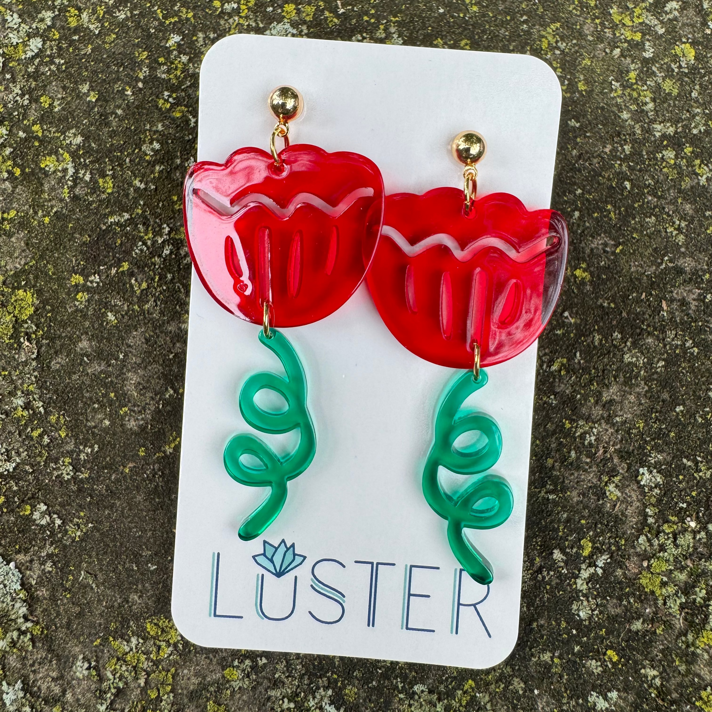 Red flower-shaped earrings with green stems on a Luster branded card against a textured stone background.
