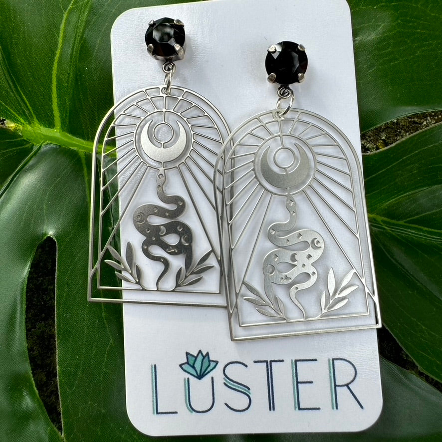 A pair of nickel-free plated steel earrings with celestial serpent designs and jet black crystal accents, displayed on a white card with the brand name 'LUSTER'.