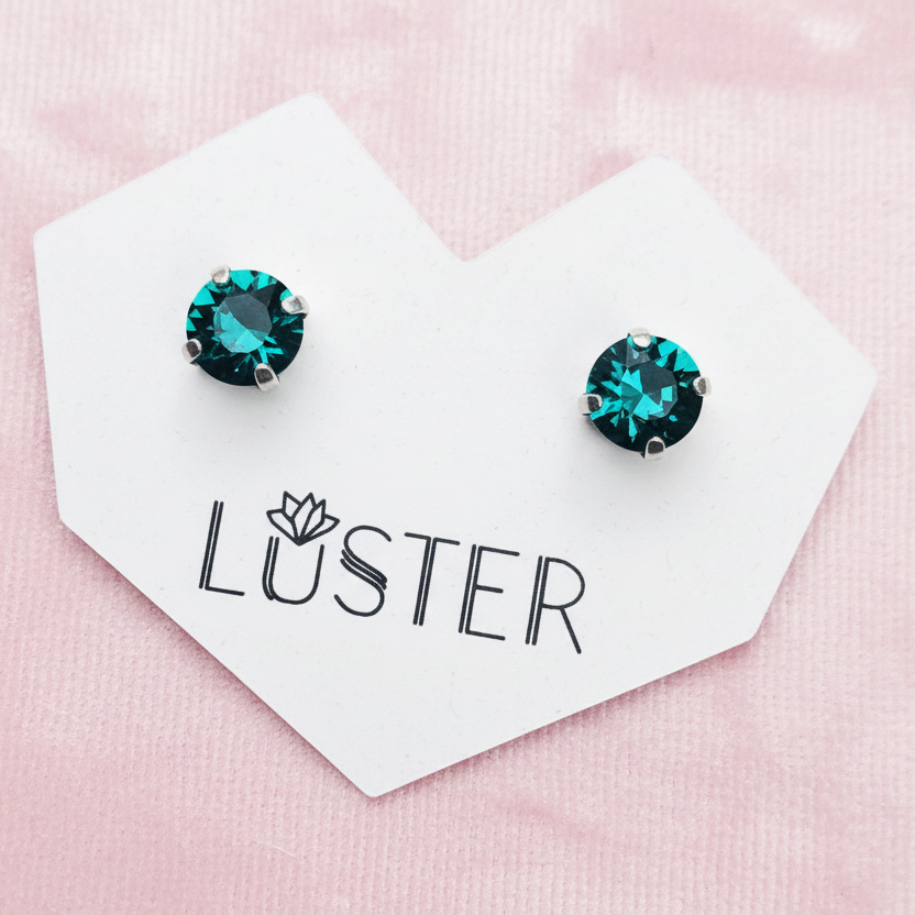 A pair of round stud earrings with deep emerald green faceted gemstones in silver settings, displayed on a white heart-shaped card printed with the brand name 'LUSTER' against a soft pink textured background.