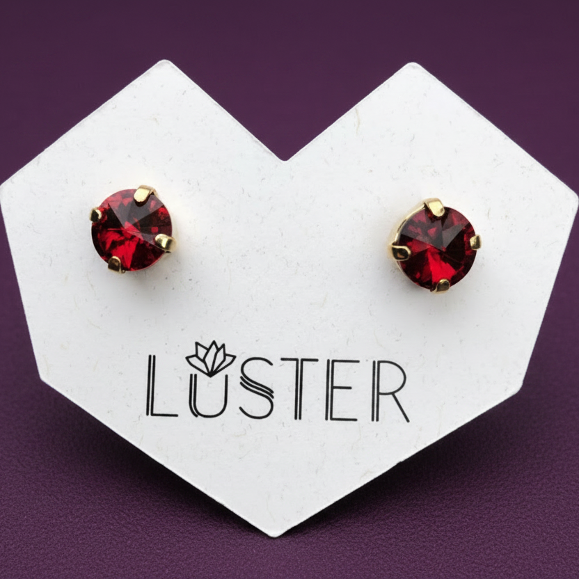 A pair of round stud earrings with deep red faceted gemstones in gold settings, displayed on a white heart-shaped card printed with the brand name 'LUSTER' against a dark purple background.