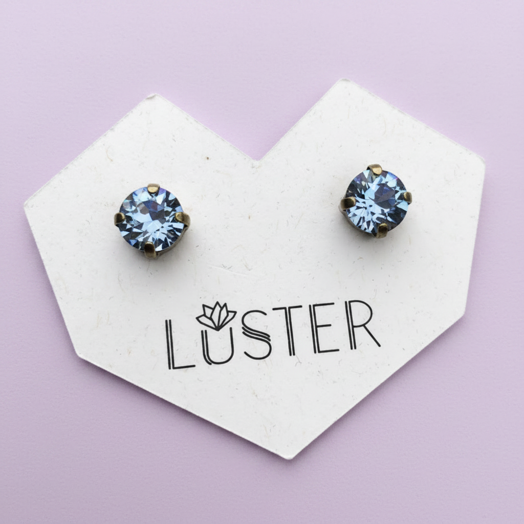A pair of round stud earrings with faceted light blue gemstones in a gold-toned setting, displayed on a white heart-shaped card printed with the brand name 'LÜSTER' against a soft lavender background.