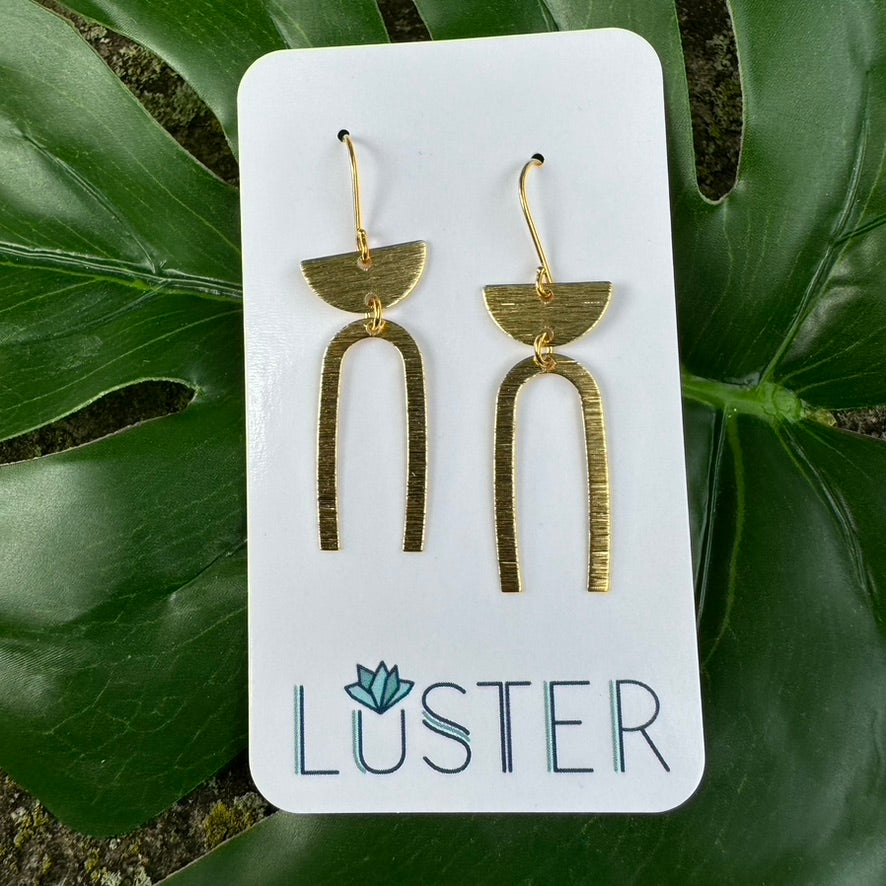 A pair of gold-colored brass earrings with a textured horseshoe shape and semicircle accents, displayed on a branded card with the word 'LUSTER'.