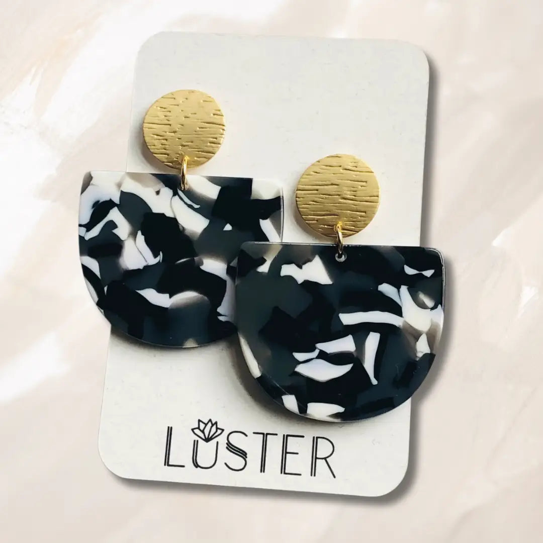 A pair of black and white swirled acrylic earrings with matte gold studs on a LUSTER branded card against an off-white background.