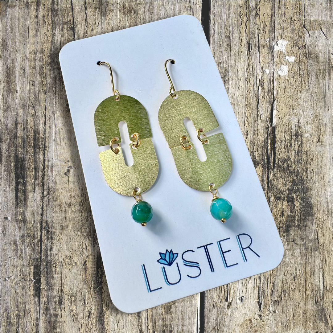 A pair of gold-colored brass dangle earrings with green faceted stone rounds, displayed on a white card with the brand name 'LUSTER' written at the bottom.