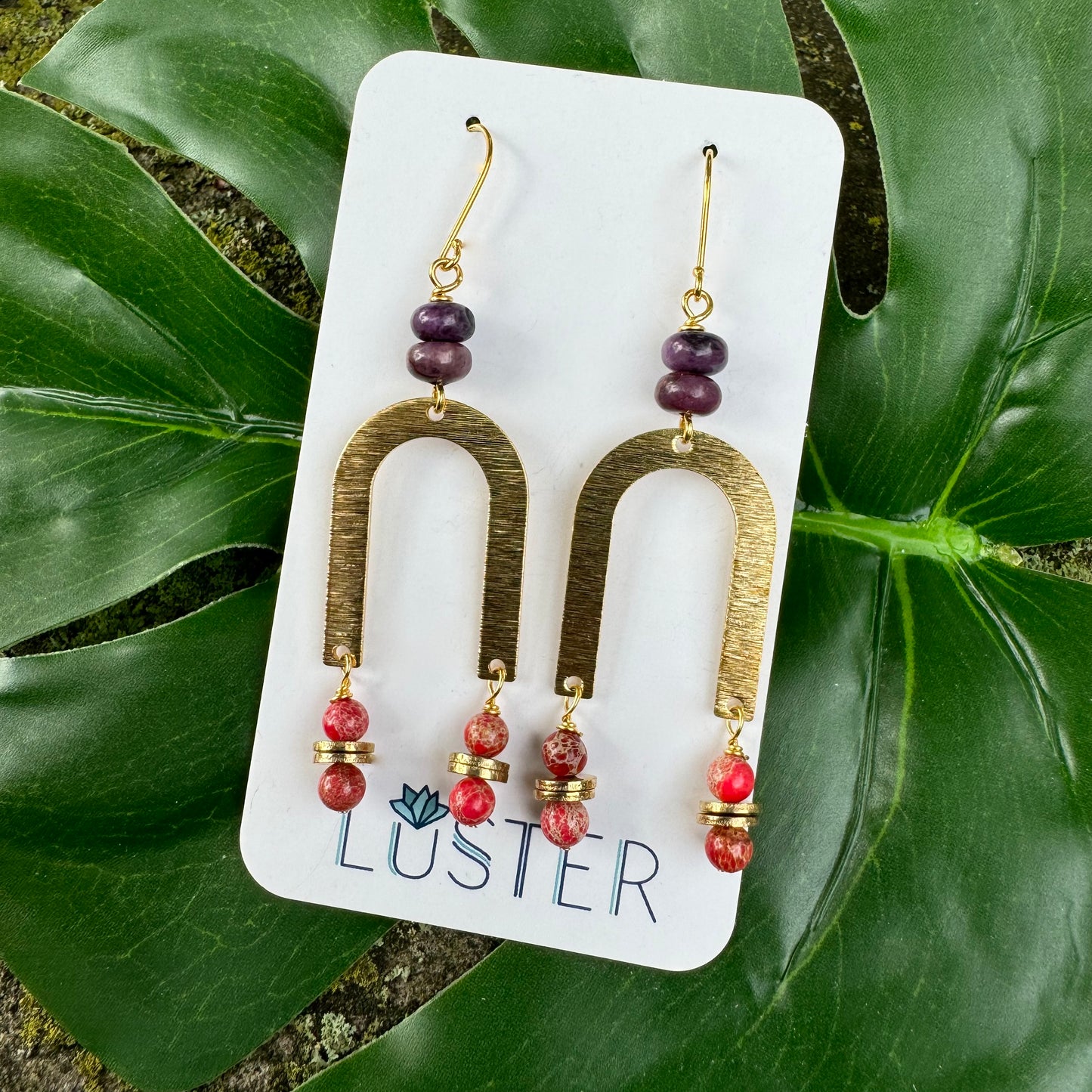 A pair of art deco style dangle earrings with orange and purple stone accents, displayed on a white card with the brand name 'luster'.