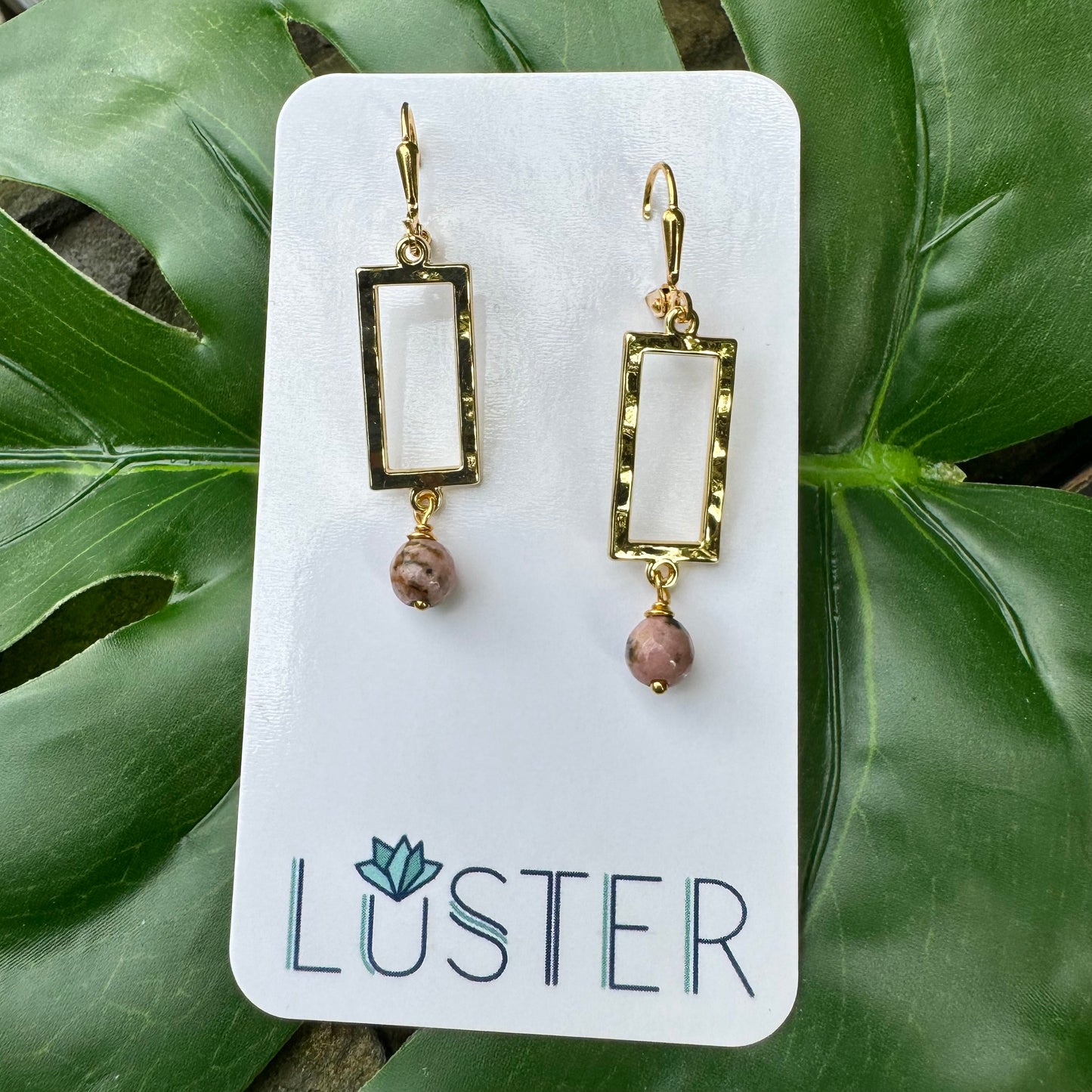 A pair of gold-plated rectangle drop earrings with rhodonite pink stone accents displayed on a white card with the brand name 'LUSTER' written on it, placed on a green leafy surface.