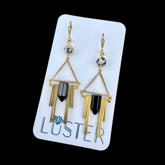 A pair of Art Deco style brass and goldstone earrings with black onyx spikes and dalmatian jasper stones, displayed on a white card with the brand name 'Luster'.