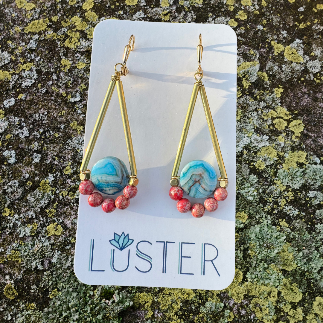 A pair of gold brass earrings with streaked teal crazy lace agate coins and red-orange impression jasper beads, displayed on a card with the brand name LUSTER.