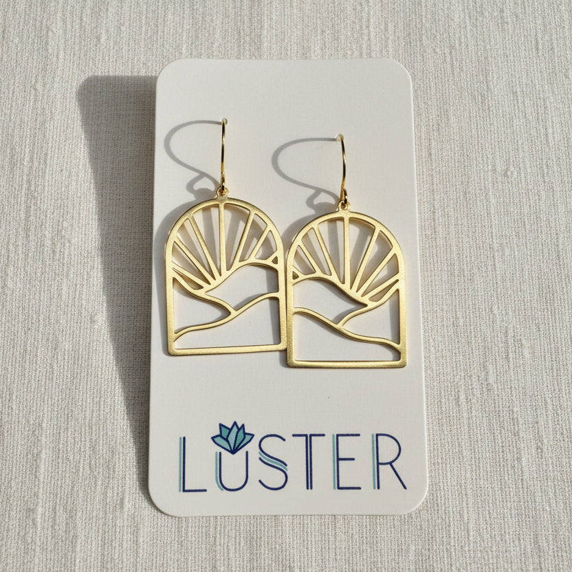 A pair of gold-tone geometric dangle earrings displayed on a white Luster brand card against a light textured fabric background.