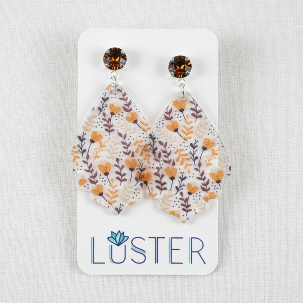 A pair of teardrop-shaped dangle earrings with a white background and a floral pattern in orange, brown, and purple, hanging from dark amber faceted studs on a white display card labeled 'LUSTER', held by a hand.