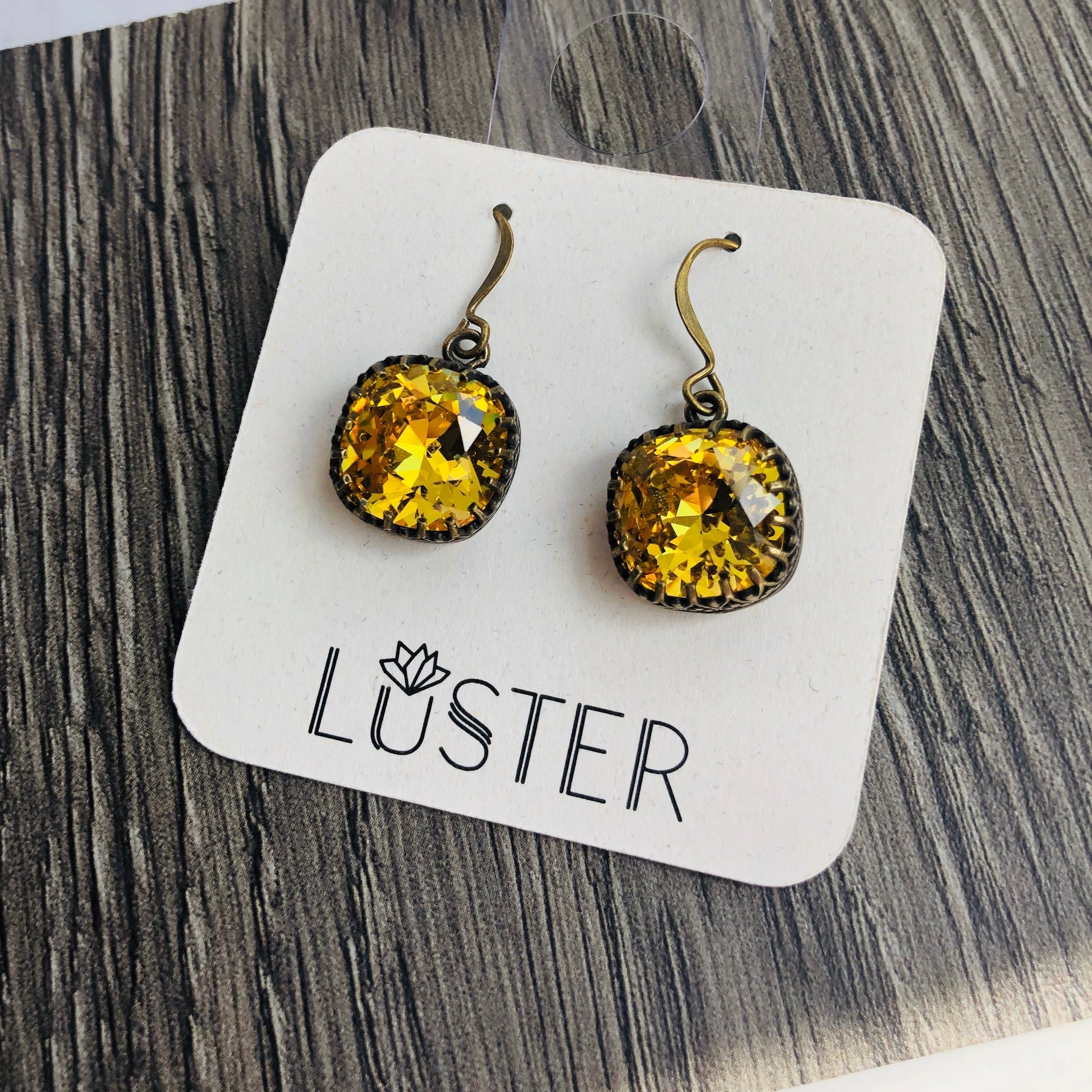 A pair of sunflower yellow cushion cut dangle earrings with antique brass bezels and French hook earwires, packaged on a card with the brand name 'Luster'.