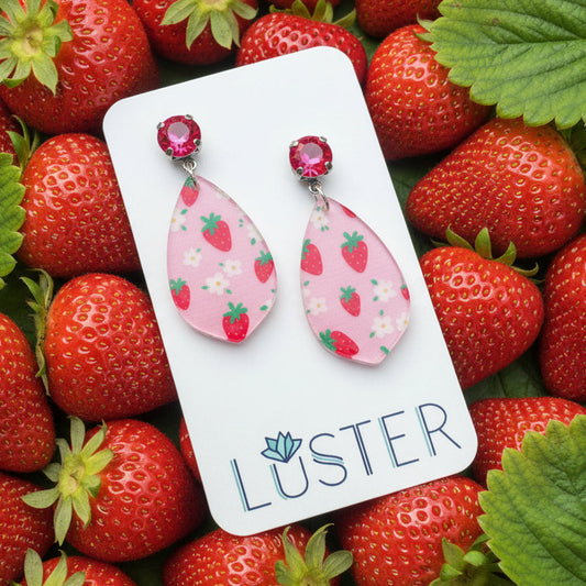 Pair of pink strawberry patterned earrings on a Luster branded card on a heap of fresh strawberries.