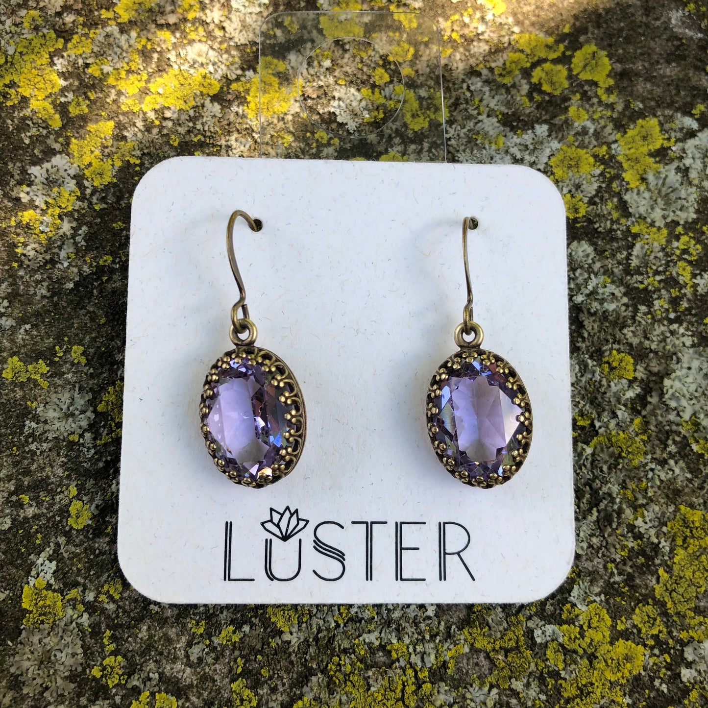 A pair of vintage-style earrings with translucent violet stones set in antique brass bezels, displayed on a white card with the brand name 'LUSTER'.