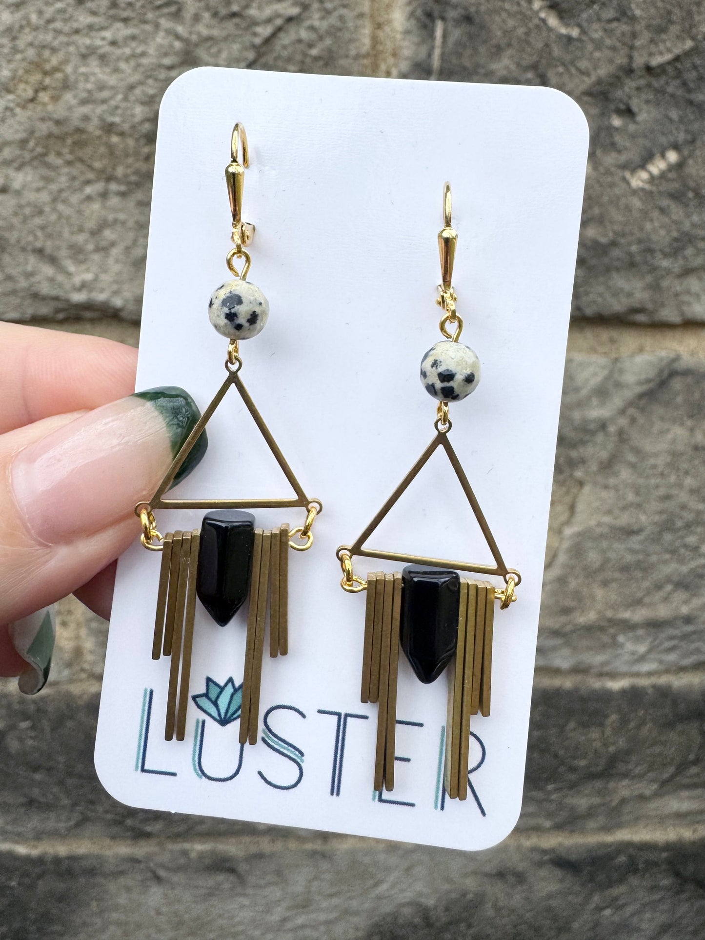 A pair of Art Deco style brass and goldstone earrings with black onyx spikes and dalmatian jasper stones, displayed on a white card with the brand name 'Luster' held up in front of a stone wall.