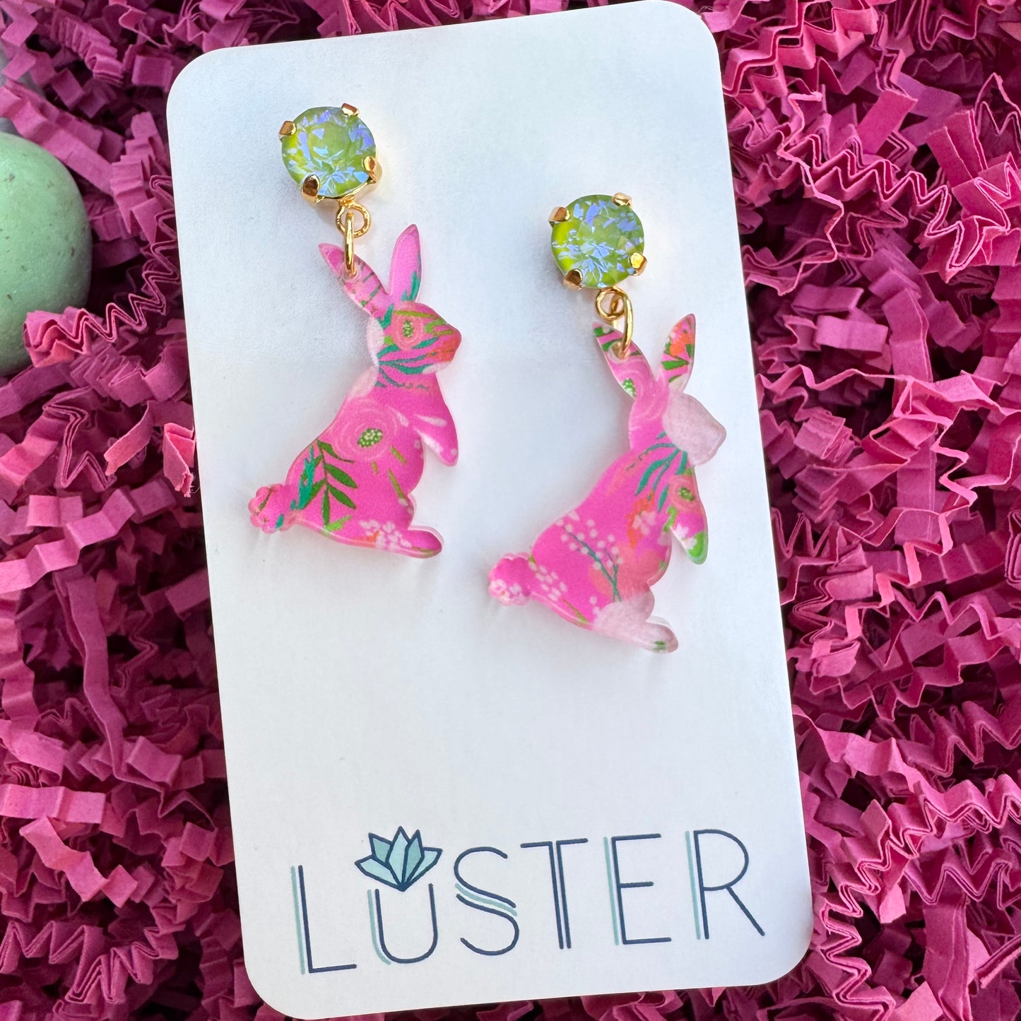 A pair of pink floral acrylic bunny earrings with sparkly lime crystal accents on a nickel-free plated brass stud, displayed on a white card with the brand name LUSTER.