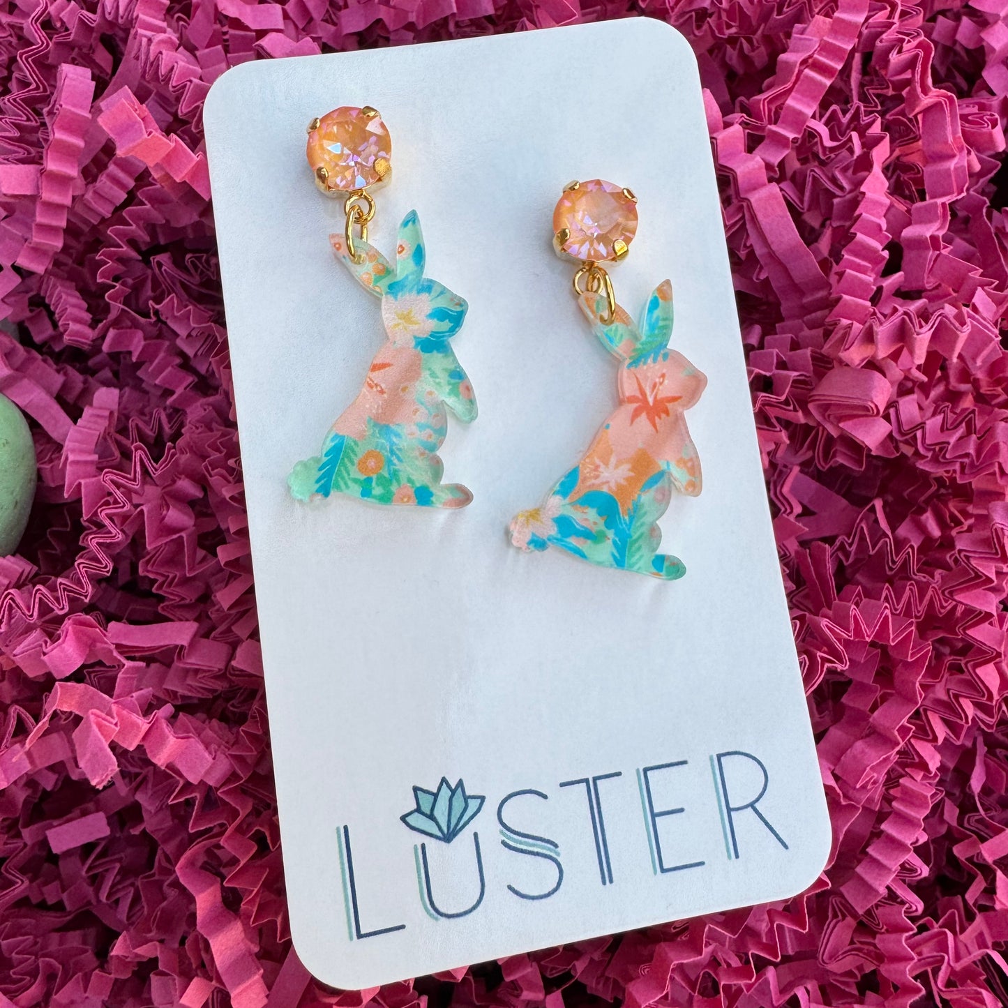 A pair of earrings designed in the shape of rabbits with floral patterns and peach crystal accents, displayed on a card with the brand name LUSTER.