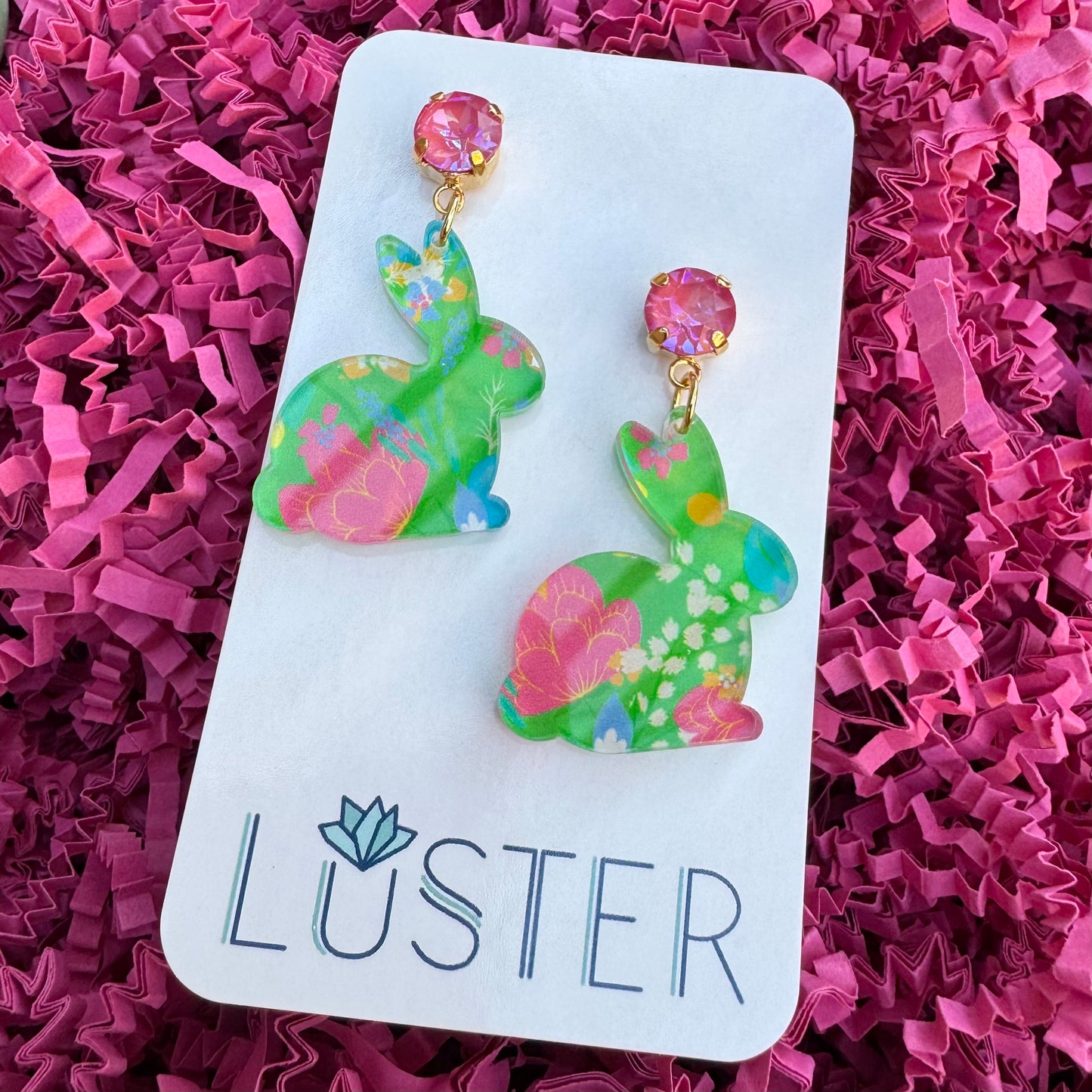 A pair of green floral acrylic earrings with pink crystal accents on a nickel-free brass stud, displayed on a white card with the brand name LUSTER written on it.