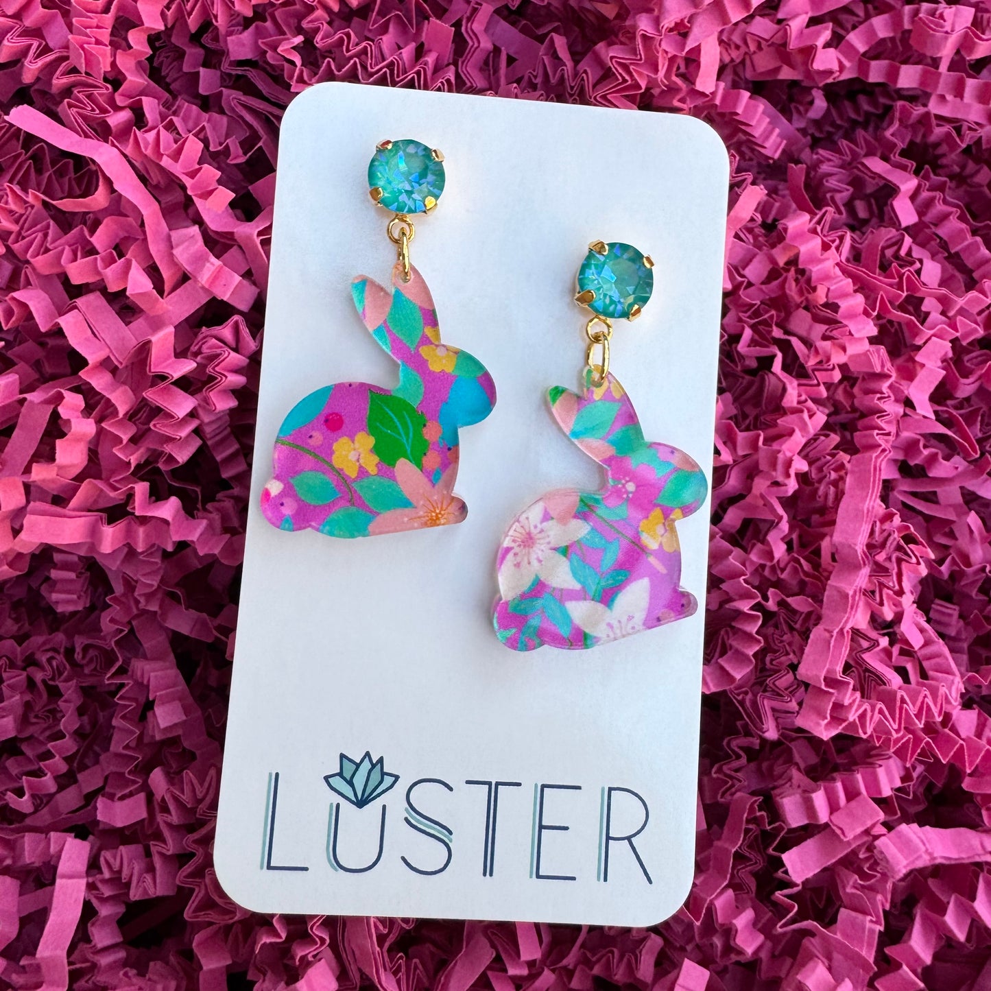 A pair of colorful acrylic bunny-shaped earrings with teal crystal accents on top, displayed on a white card with the brand name LUSTER written on it, against a background of pink crinkle paper.