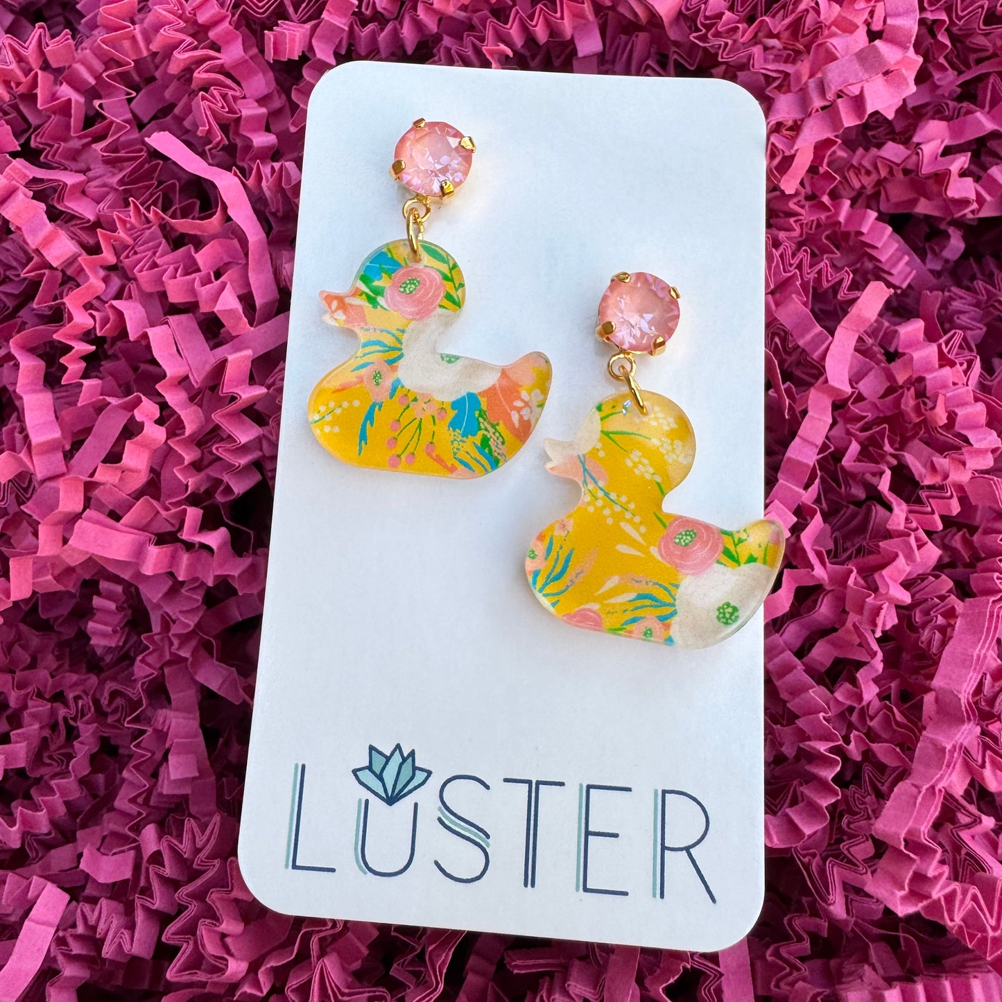 A pair of colorful acrylic duck earrings with pink crystal accents on a nickel-free brass stud, displayed on a pink crinkle paper with a brand card reading 'LUSTER'.