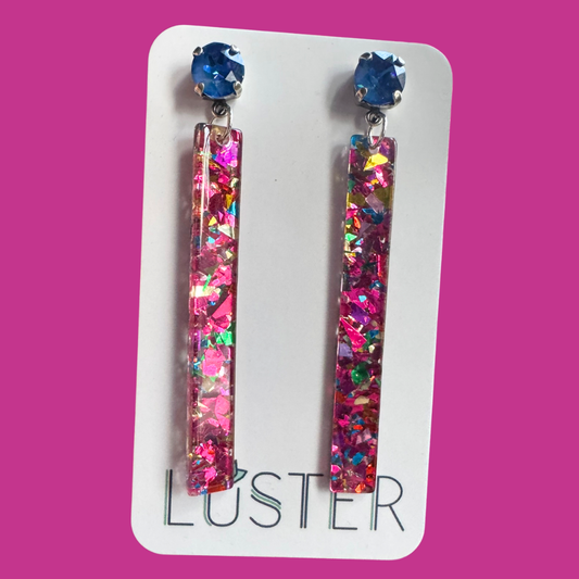 Colorful glittery earrings on a Luster branded card against a pink background