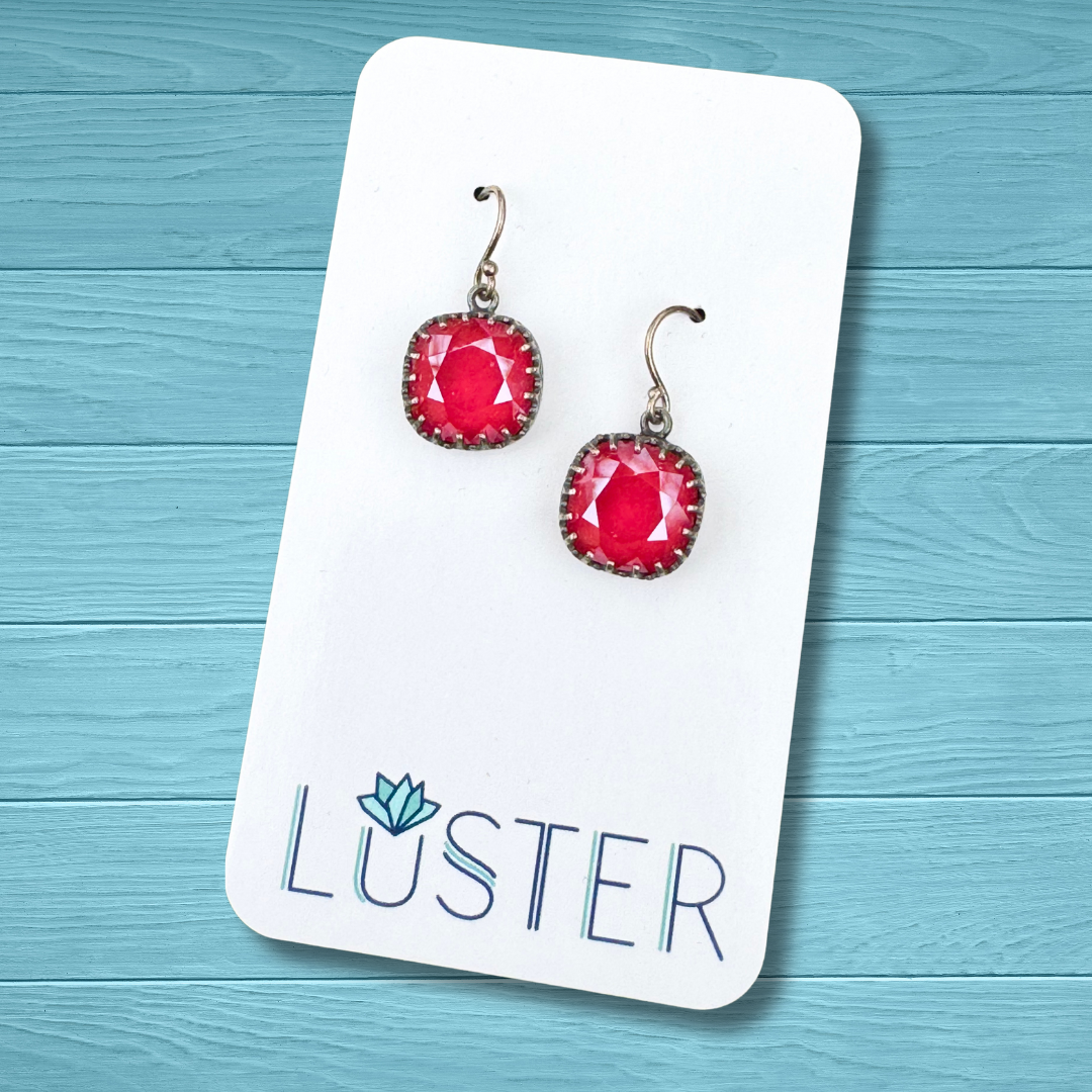 A pair of cushion cut dangle earrings with royal red crystals, set in antique silver bezels with sterling silver French hook earwires, displayed on a white card with the brand name LUSTER.