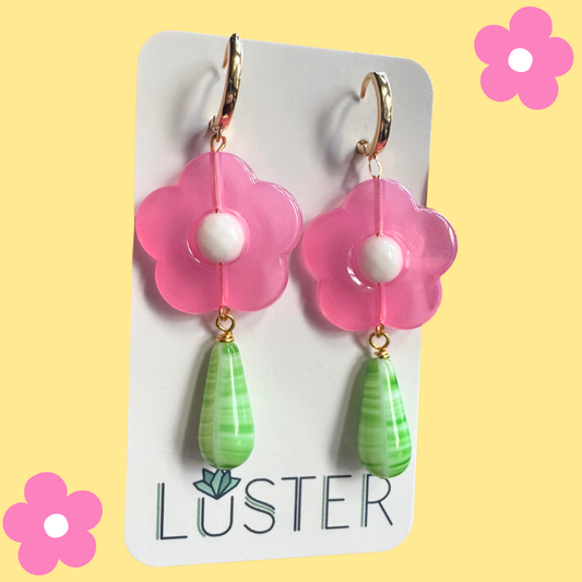 Flower-shaped earrings with pink flowers and green teardrop charms on a Luster branded card against a yellow background.