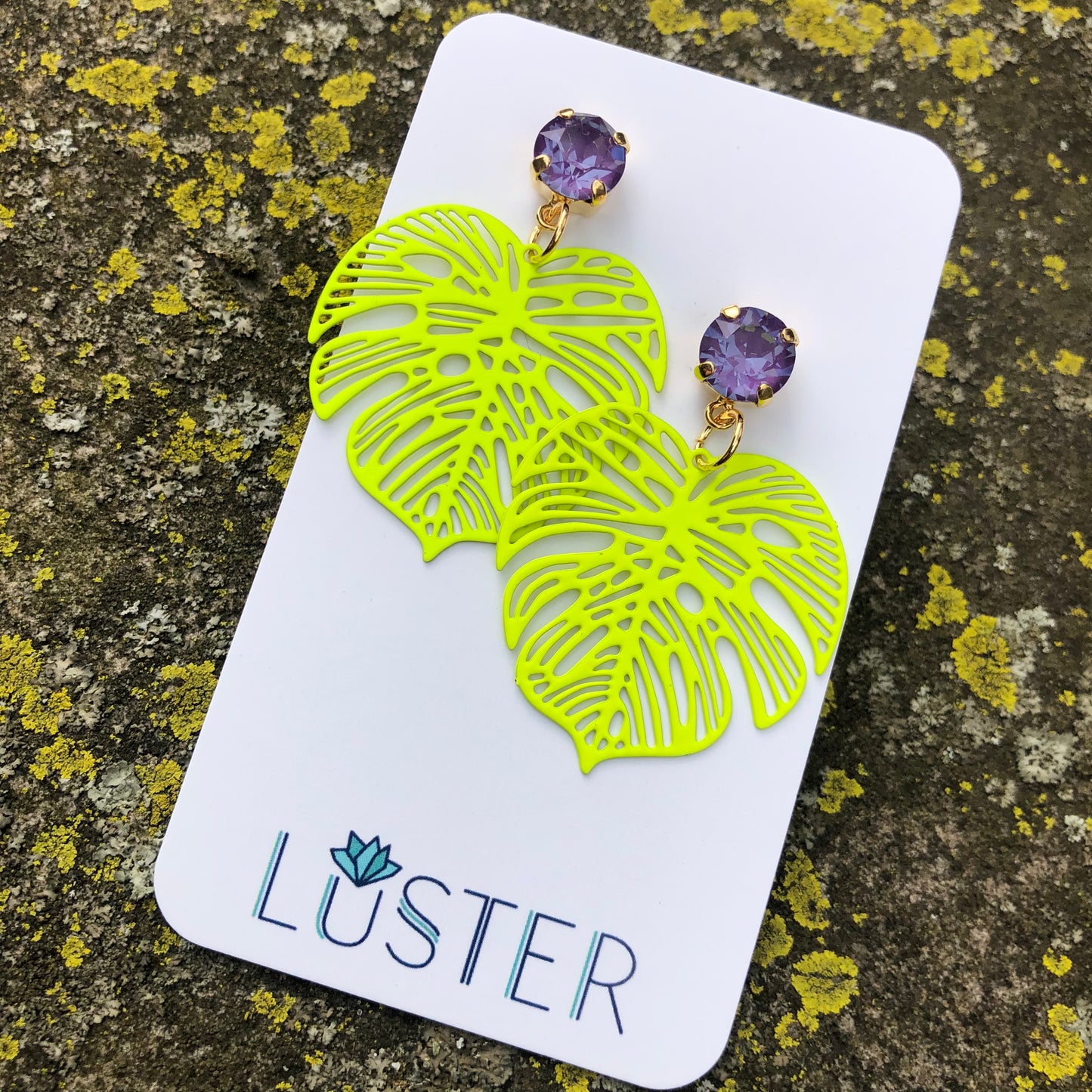 A pair of bright yellow-green enamel leaf-shaped earrings with purple crystal accents displayed on a branded card.