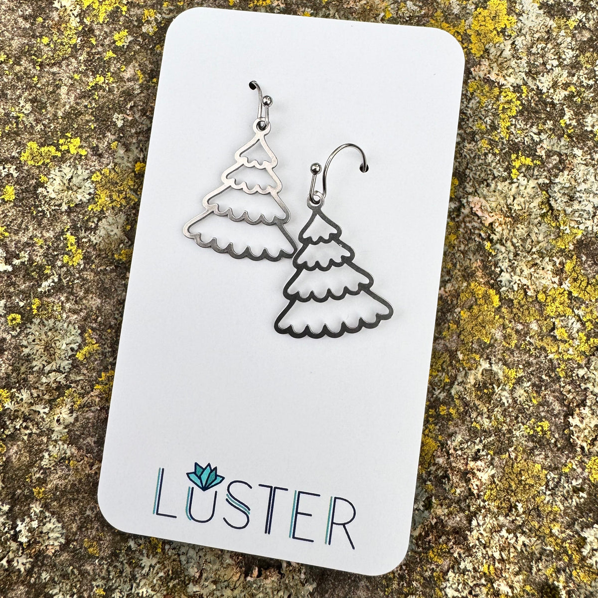 A pair of silver-colored stainless steel earrings in the shape of snowy trees, displayed on a white card with the brand name 'LUSTER' written at the bottom.