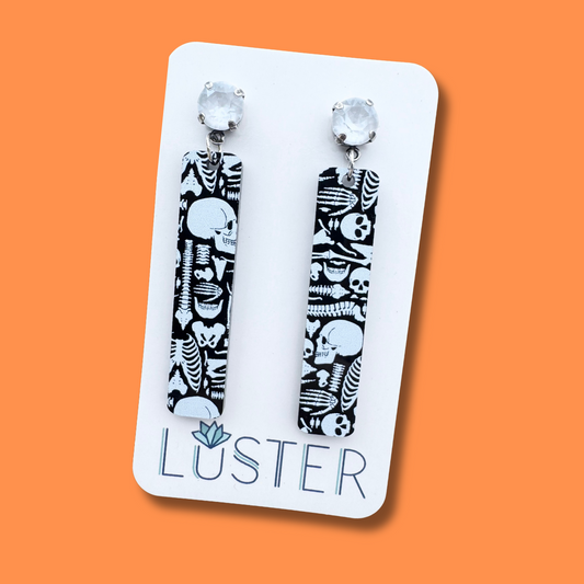 Skateboard-shaped earrings with skull design on a Luster branded card against an orange background