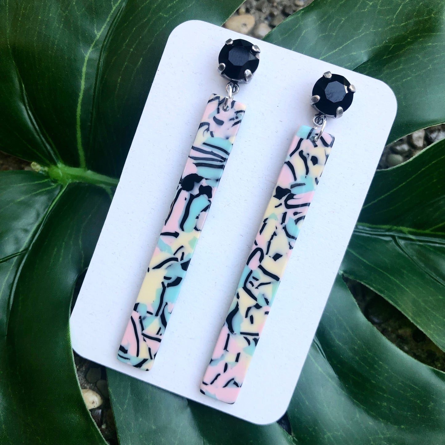 A pair of acrylic stick earrings with jet crystal accents, displayed on a white holder, placed against a green leafy background.