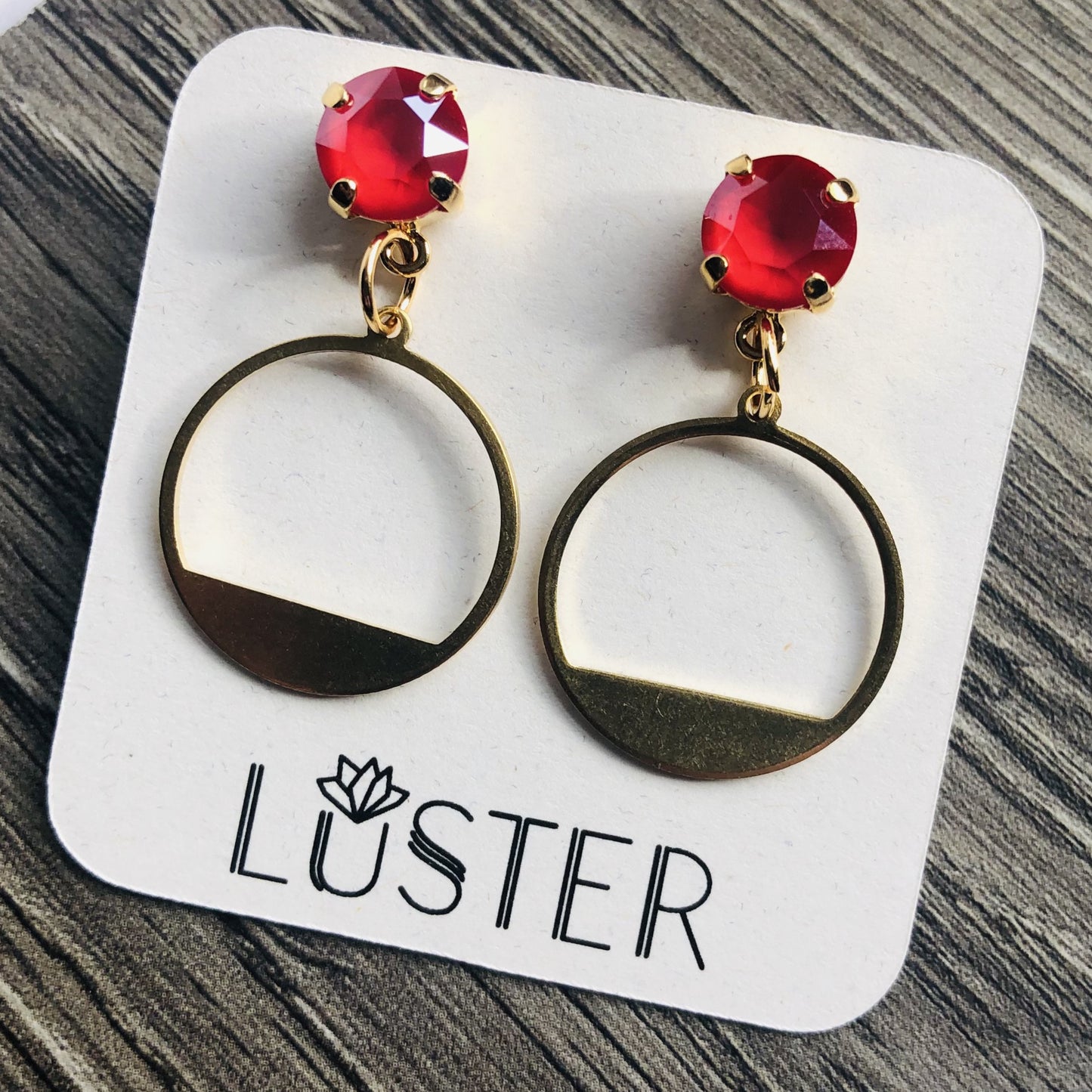 A pair of gold-colored round drop earrings with red crystal accents, displayed on a card with the brand name 'LUSTER'.