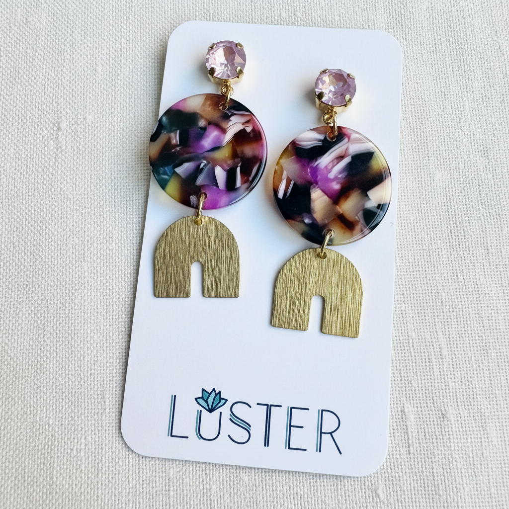 A pair of drop earrings displayed on a white Luster brand card against a light beige fabric background. The earrings feature round marbled resin discs in purple, pink, black, and gold tones, hanging from gold-toned posts with a faceted pink gemstone and a textured gold arch charm.
