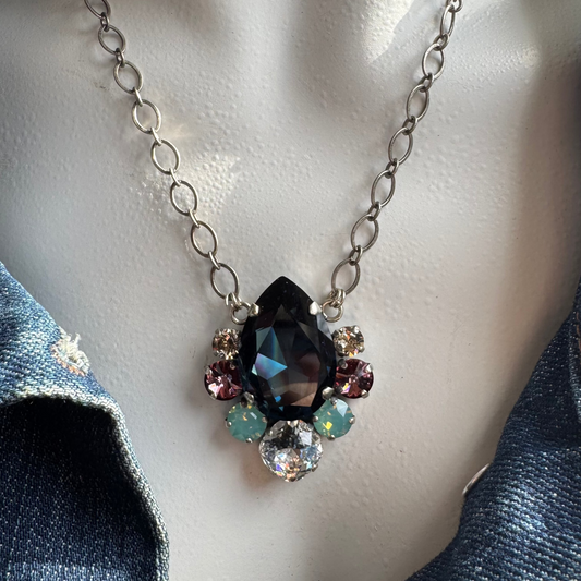 A bold teardrop pendant necklace with navy blue, pacific opal, and antique pink shades, displayed on a mannequin wearing a denim jacket.