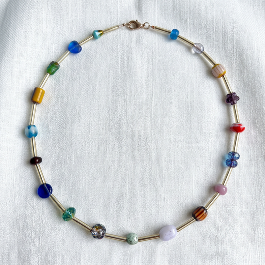 A beaded necklace with gold-toned metal links and multicolored glass beads, arranged in a circle on a white textured fabric background.