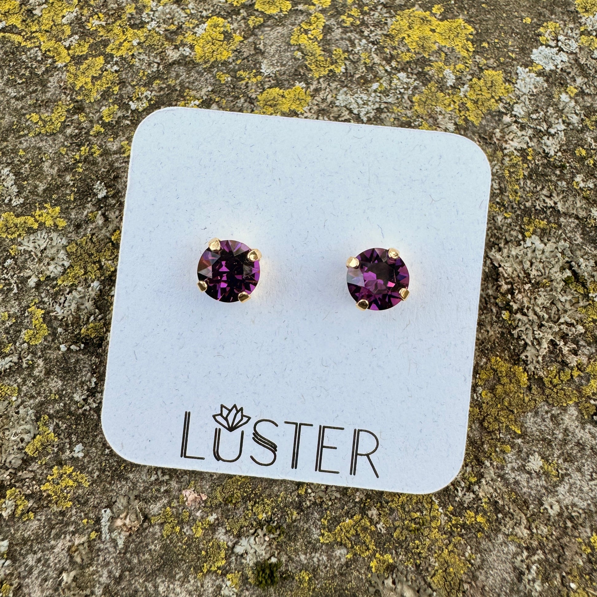 Purple round stud earrings on a Luster branded card against a textured stone background
