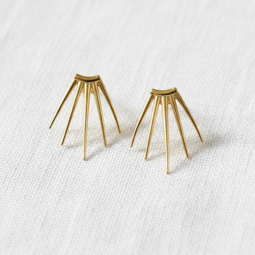A pair of gold geometric stud earrings with a starburst or sunburst design, resting on a textured white linen surface.