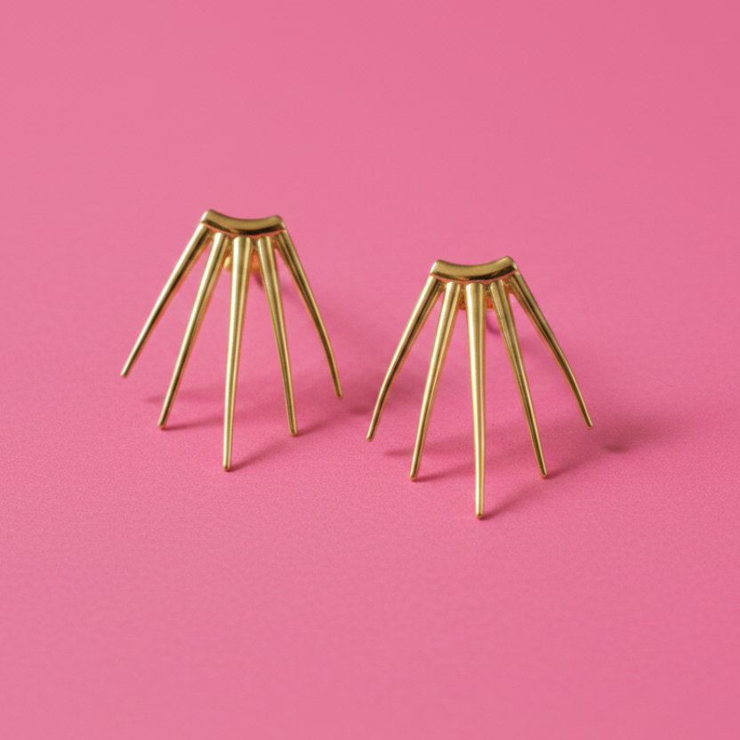 A pair of gold-toned spider-themed stud earrings with multiple thin legs, displayed on a solid pink background.
