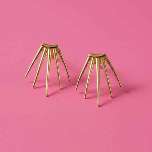 A pair of gold-toned spider-themed stud earrings with multiple thin legs, displayed on a solid pink background.