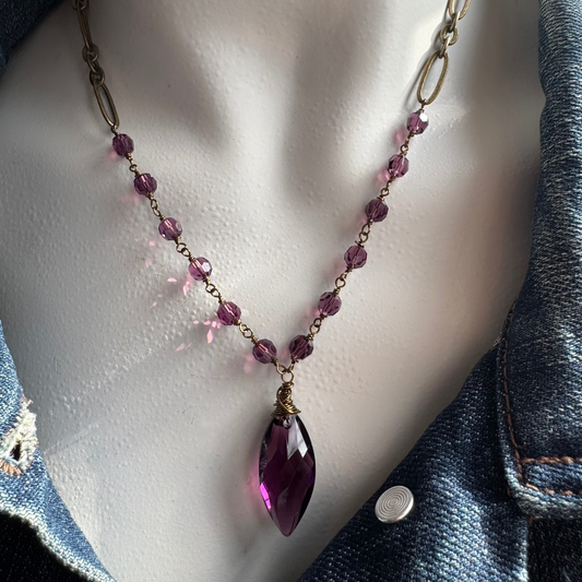 A faceted Swarovski amethyst hand-wrapped pendant necklace with round crystal accents and an adjustable antique brass chain, displayed on a mannequin wearing a denim jacket.