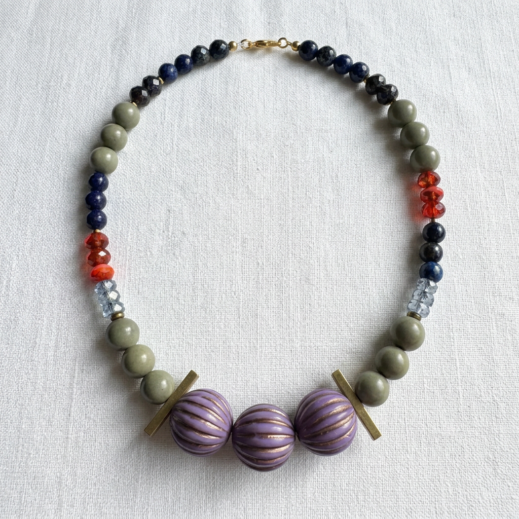 A handmade beaded necklace with olive green, navy blue, and red beads, featuring three large purple striped glass beads at the center and gold-tone metal accents, displayed on a textured white fabric background.