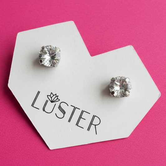 A pair of silver-toned stud earrings with clear faceted gemstones, displayed on a white hexagonal card printed with the word 'LUSTER' against a bright pink background.