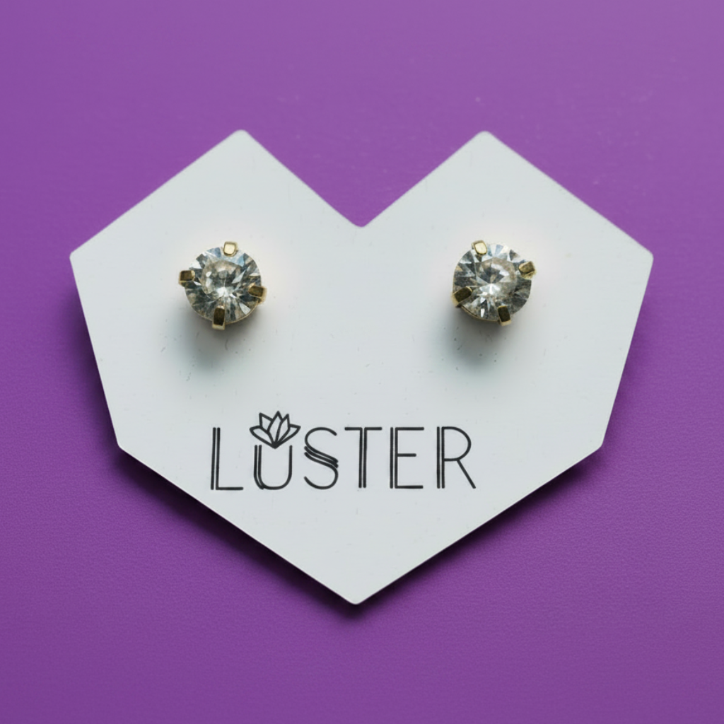 A pair of gold stud earrings with clear faceted gemstones, displayed on a white heart-shaped card printed with the brand name 'LUSTER' against a solid purple background.