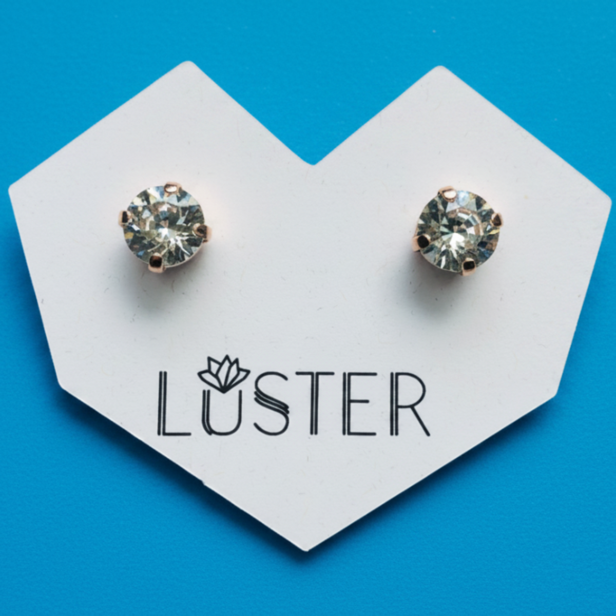 A pair of round bezel-set clear gemstone stud earrings displayed on a white heart-shaped card printed with the brand name 'LUSTER', set against a bright blue background.