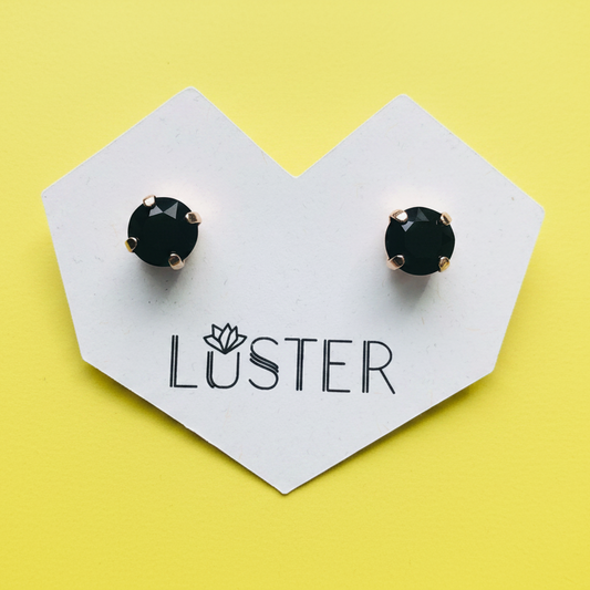 A pair of round black faceted stud earrings with gold-toned prongs, displayed on a white heart-shaped card printed with the brand name 'LUSTER' against a bright yellow background.