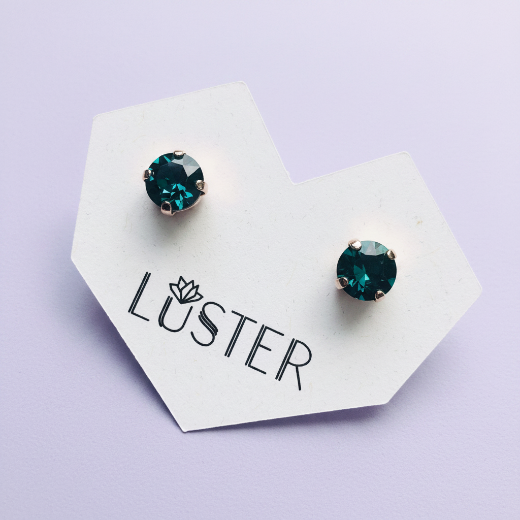 A pair of round stud earrings with deep emerald green faceted gemstones in silver-toned settings, displayed on a white branded card that reads 'LUSTER' against a light lavender background.