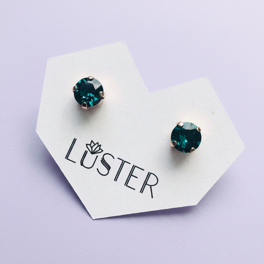 A pair of round stud earrings with deep emerald green faceted gemstones in silver-toned settings, displayed on a white branded card that reads 'LUSTER' against a light lavender background.