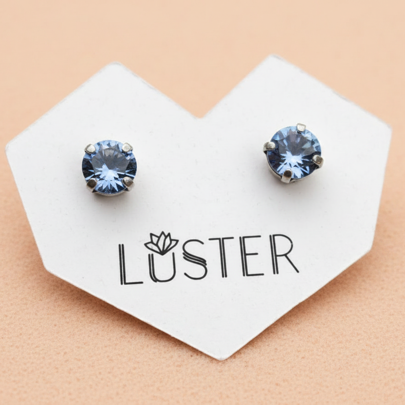 A pair of round stud earrings with light blue faceted gemstones in silver-toned prong settings, displayed on a white heart-shaped card printed with the brand name 'LUSTER' against a peach background.