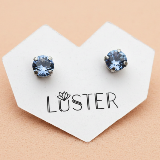 A pair of round stud earrings with light blue faceted gemstones in silver-toned prong settings, displayed on a white heart-shaped card printed with the brand name 'LUSTER' against a peach background.