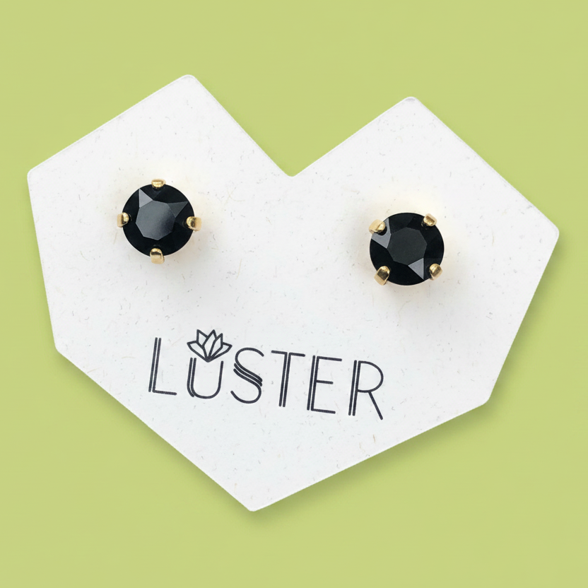 A pair of round black faceted stud earrings with gold prongs, displayed on a white branded Løster card against a light green background.