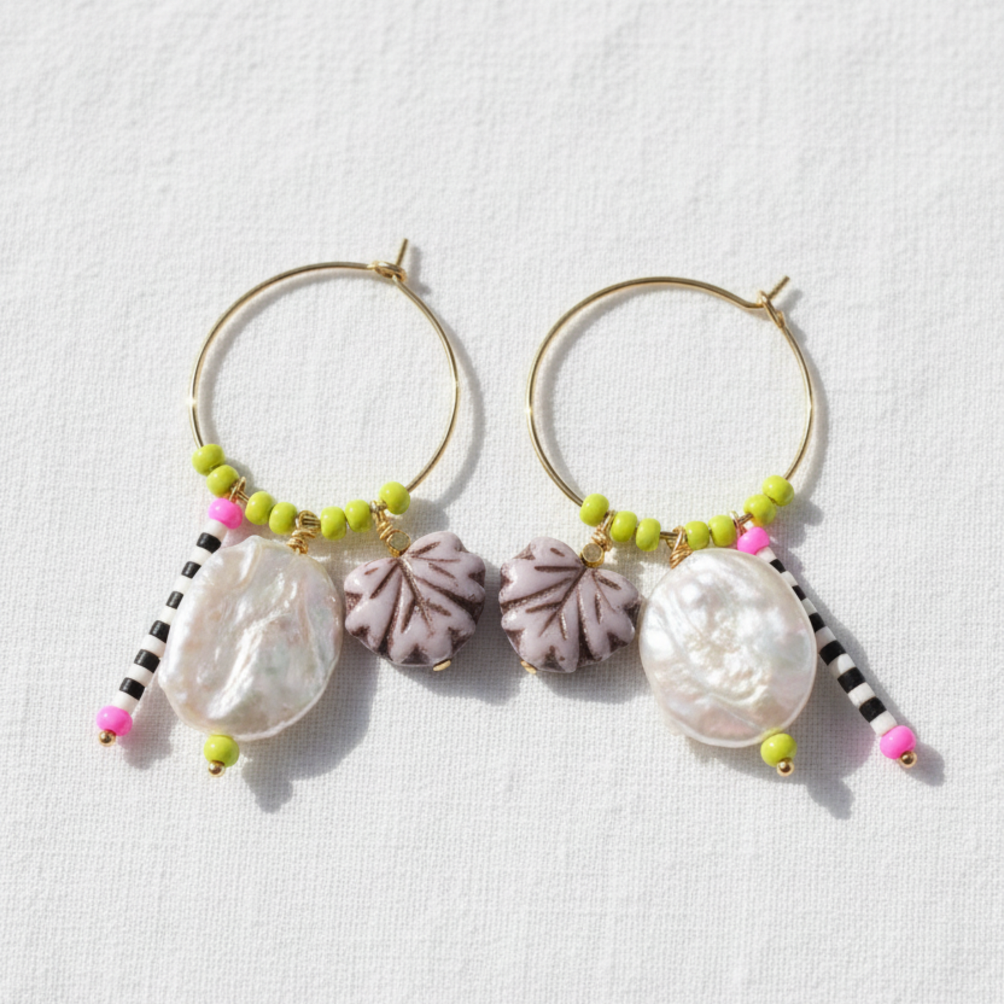 A pair of gold hoop earrings with dangling baroque shell charms, purple textured flower beads, lime green beads, and black-and-white beaded tassels, displayed on a textured white background.